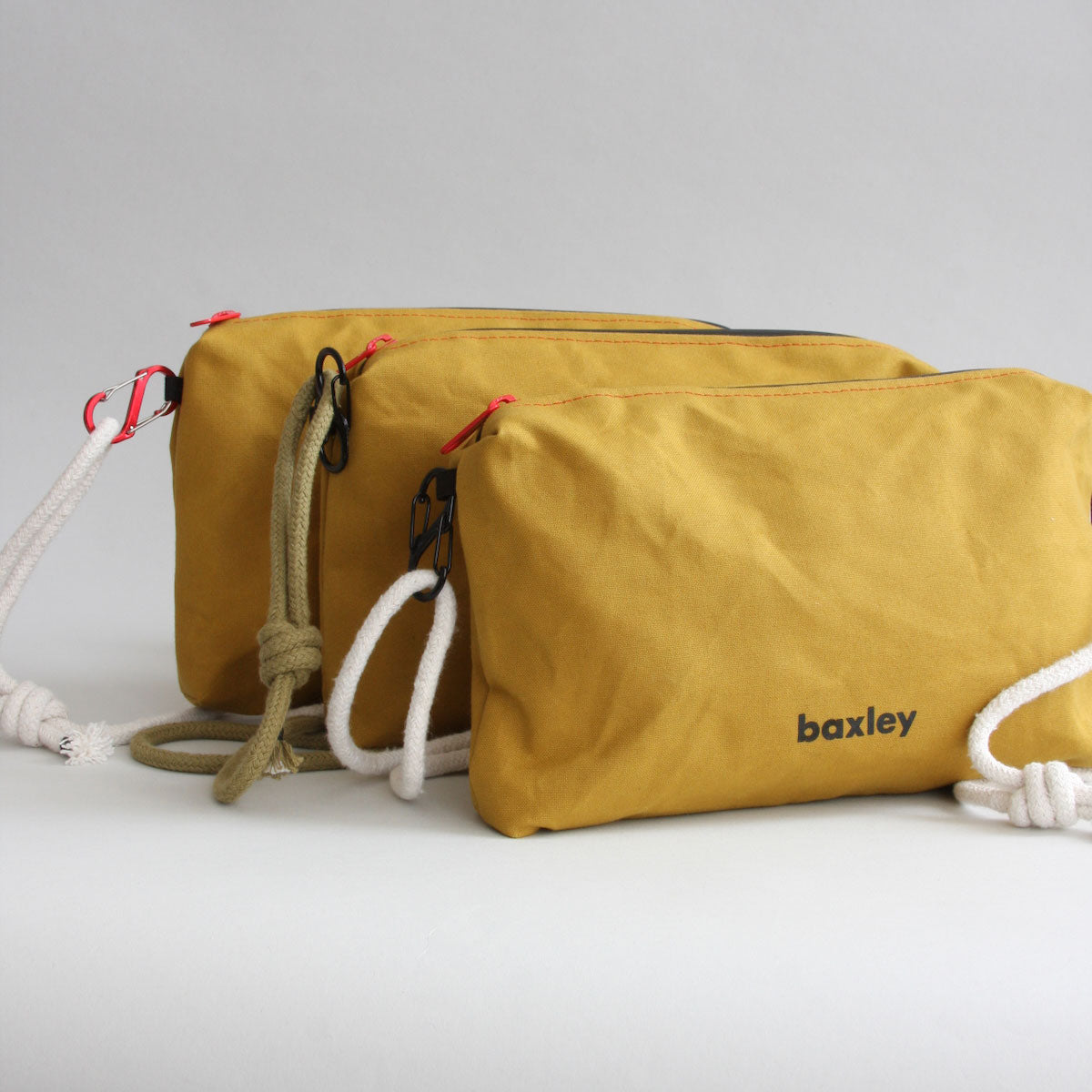 Baxley's Effie crossbody pouch shown with three variations. All canvas is cumin, two ropes are white, one rope is green. Two of the carbiners are black, one is red. The zip sliders are all red.