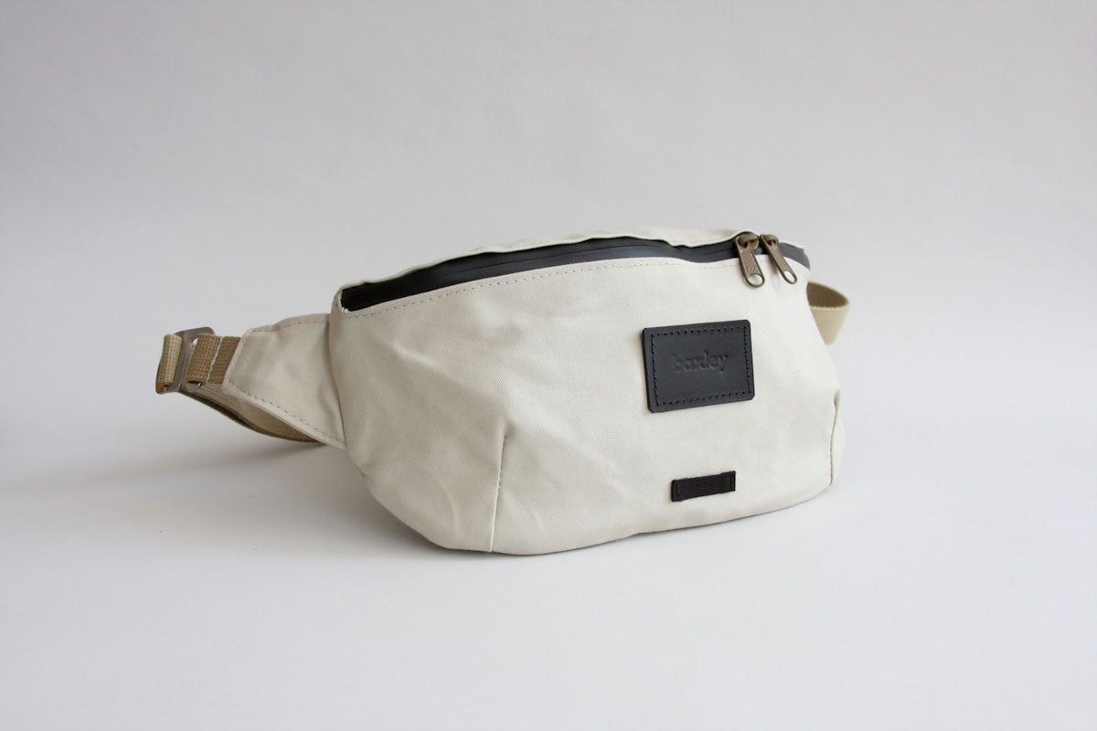 An angled front view of Otis the sling. The bag is a chalk white in colour, with beige webbing, black zipper and black leather badge. The zip pulls are the same beige as the webbing. There is a silver metal g-hook clasp closing the strap.