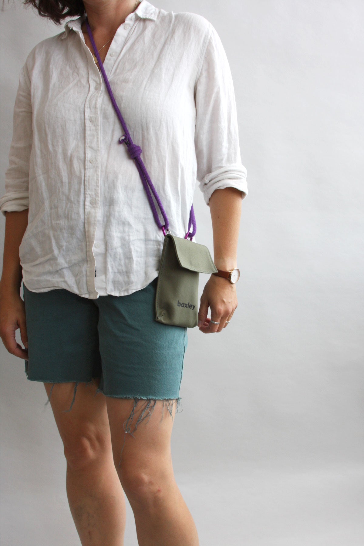 Pip the pocket worn cross-body on a model. The bag is sage green, has a screen-printed logo, and a purple rope strap. The model is in a studio setting, wearing informal clothing.
