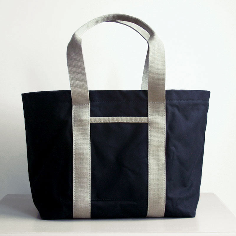 Front view of Hank the tote from Baxley. Shown in black with beige webbing, with a front pocket visible.