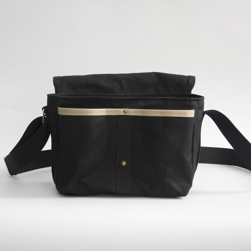 A front view of Gertie the crossbody satchel from Baxley in black, with flap in open position. A beige ridge of webbing indicates two front pockets.
