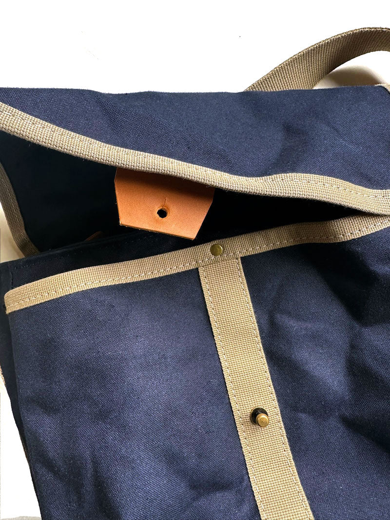 A close-up image of a midnight blue version of Gertie the crossbody satchel. A brown leather closure tab is visible, as is beige accent webbing.