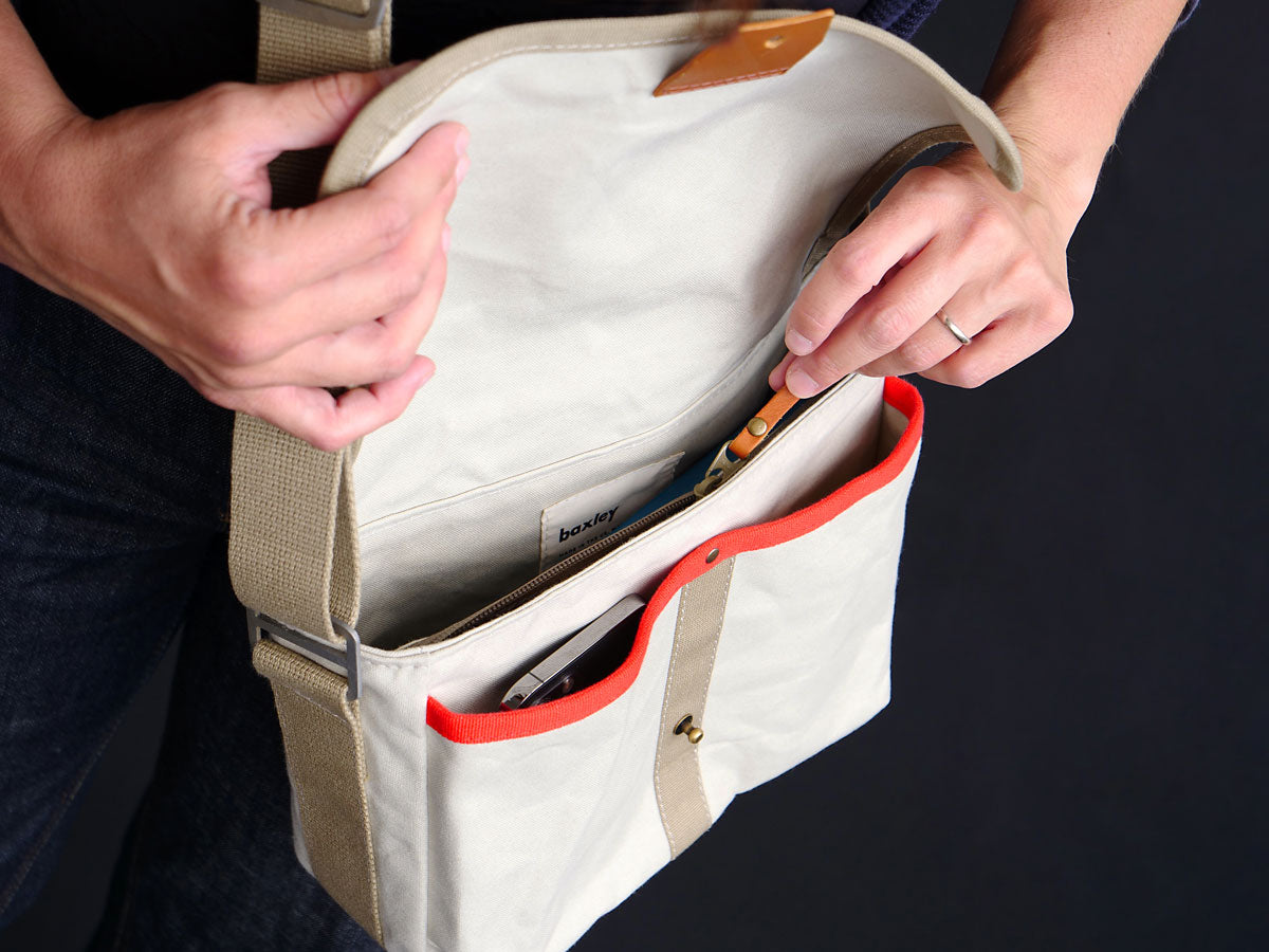Interior action pic of a bag being zipped open. The bag is a chalk white Gertie the crossbody satchel from Baxley. It has two front pockets beneath the open flap, one of which is shown holding a mobile phone.