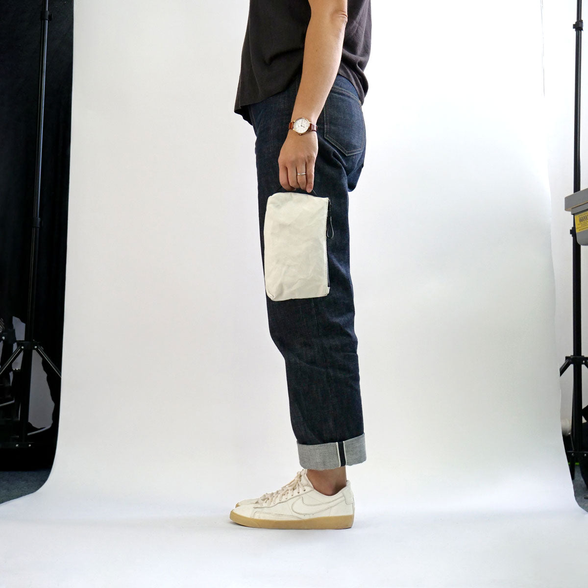 Reggie the pouch by Baxley shown in chalk white. It is being held by a side handle by a female model in a studio setting. The model is wearing jeans a t-shirt and trainers.