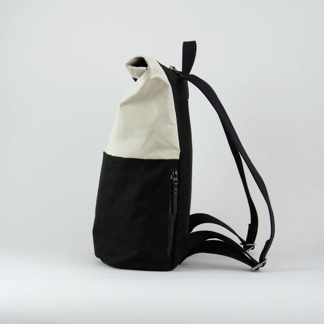 Side view of Max the backpack. A rolltop backpack made of waxed canvas in chalk white and black, with a side zipped pocket visible for storage of phone or keys.